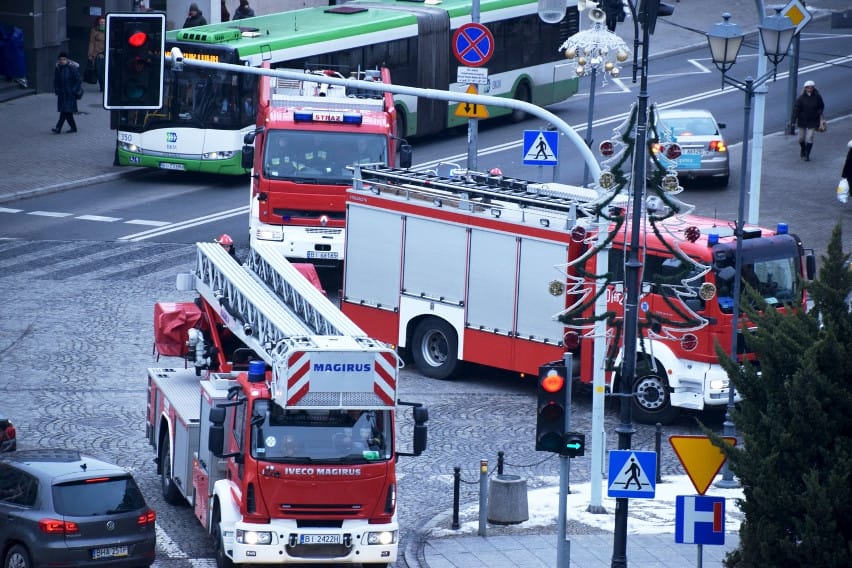 Scena przedstawiająca wozy strażackie i ewakuację ludzi przed hotelem, symbolizująca fałszywy alarm pożarowy.